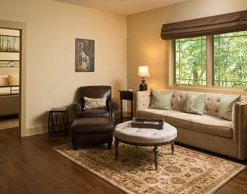 Brown Leather chair with ottoman, light tan couch with a round fabric coffee table and windows with a view of the mountains. Hard wood floors with parsley center rug